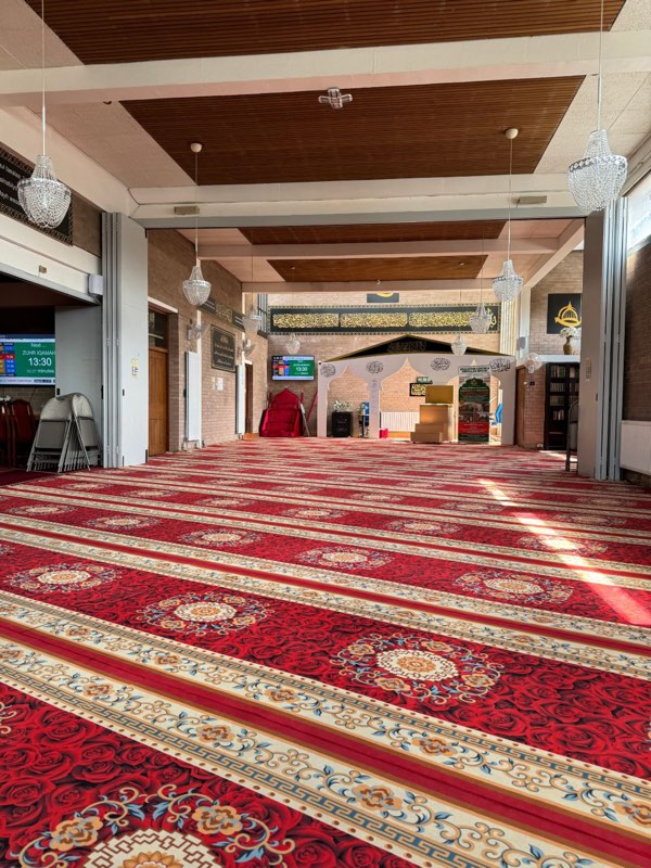 The prayer hall: rows of red patterned prayer carpet, crystal chandeliers, gold calligraphy along the far wall, and the white arched mihrab with the appeal banner standing beside it.